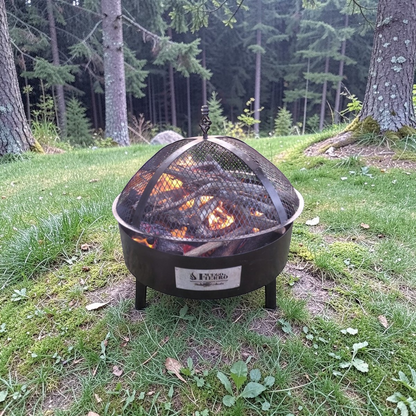 Fuego & Fierro KANSAS black metal fire pit with burning logs and mesh lid, set in a grassy backyard.